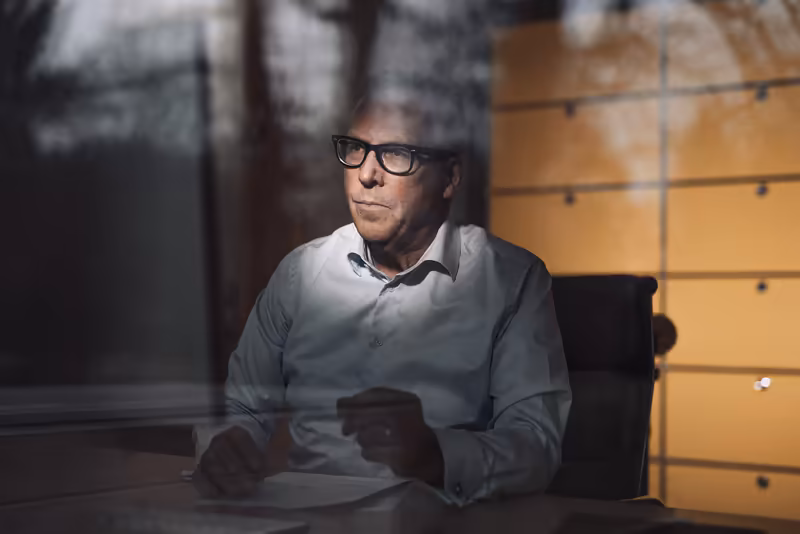 Mann mit Brille am Schreibtisch im modernen Büro, konzentrierter Blick zur Seite, weiße Hemd, warme Beleuchtung