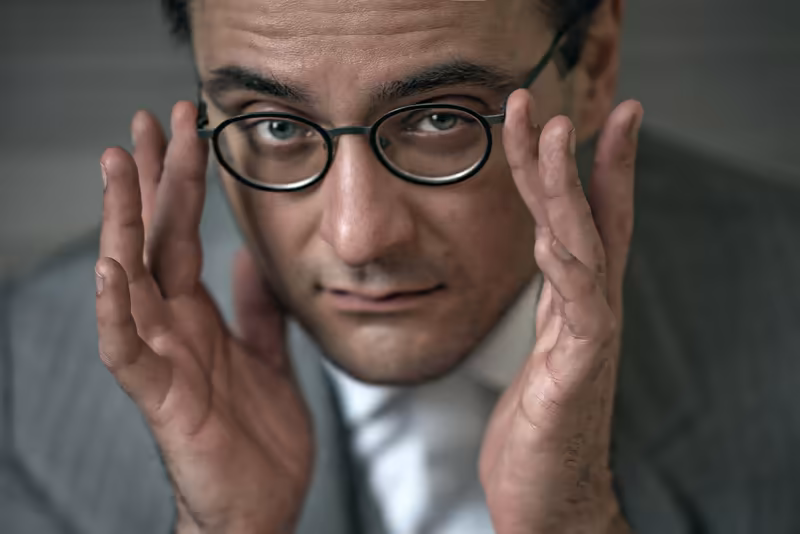 Mann mit Brille in Businesskleidung, beide Hände an den Wangen, skeptischer Gesichtsausdruck