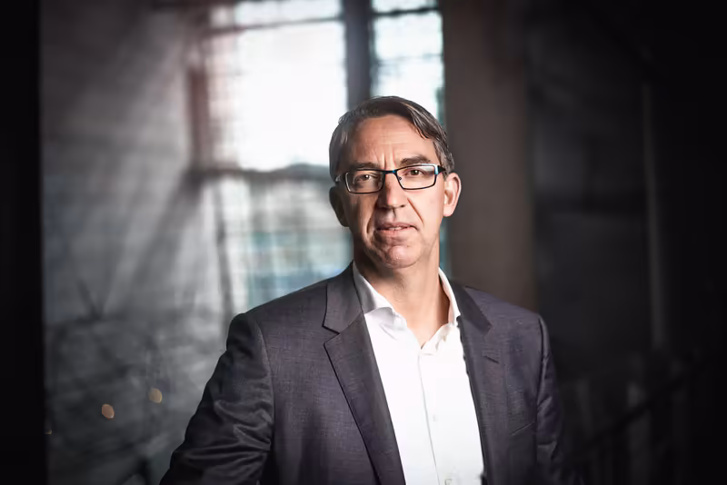 Mann mit Brille in dunkelblauem Blazer und weißem Hemd vor Fenster mit Industriekulisse, professionelles Porträt