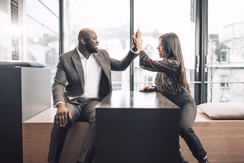 Zwei Geschäftspersonen geben sich High-Five an modernem Schreibtisch in Büro mit Skyline-Blick, erfolgreiche Zusammenarbeit