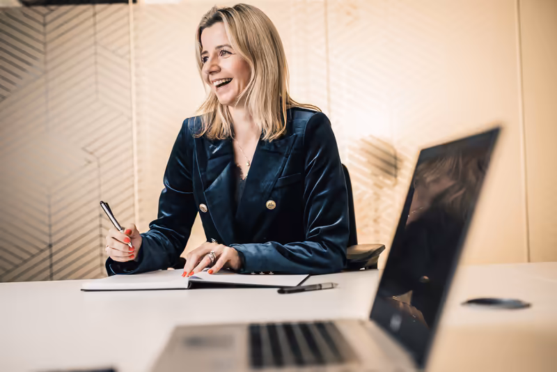 Frau in dunklem Blazer schreibt lächelnd am Schreibtisch, Laptop und Monitor im modernen Büro