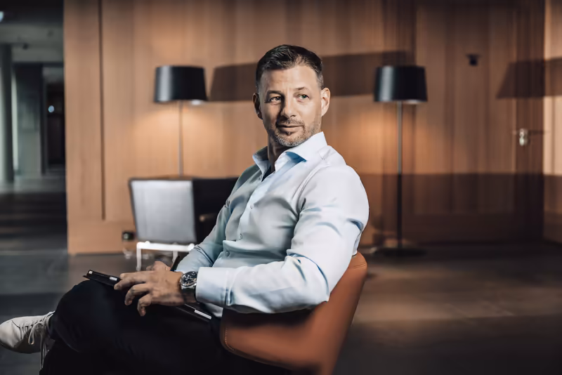 Mann in weißem Hemd sitzt in modernem Büro mit Holzwand, blickt zur Seite, professionelles Portrait