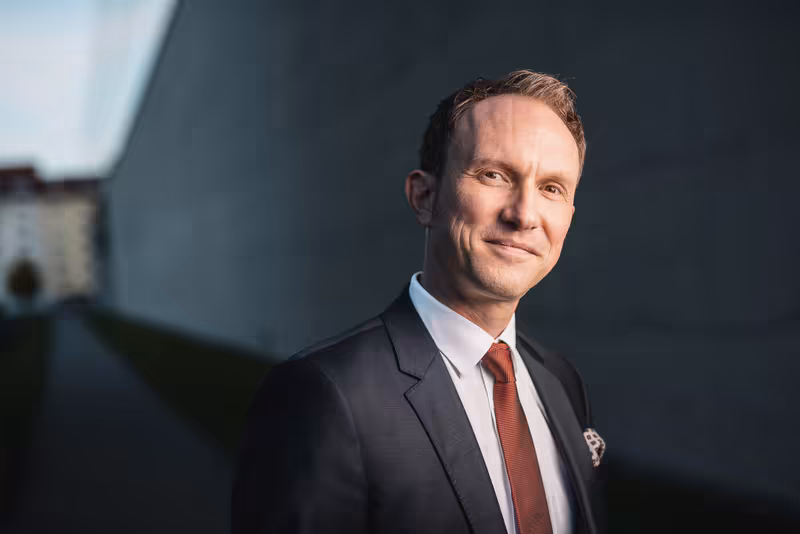 Mann im dunklen Anzug mit roter Krawatte vor grauer Architektur, professionelles Porträt mit selbstsicherem Blick