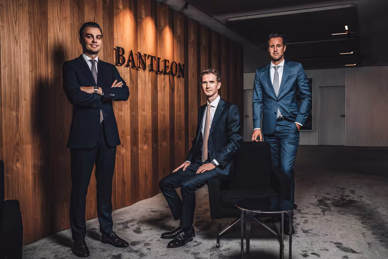 Drei Geschäftsmänner in Anzügen in modernem Büro vor Holzwand mit Bantleon-Logo, professionelles Business-Portrait