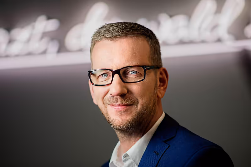 Mann mit Brille im dunkelblauen Blazer, selbstbewusstes Porträt vor unscharfem modernem Hintergrund