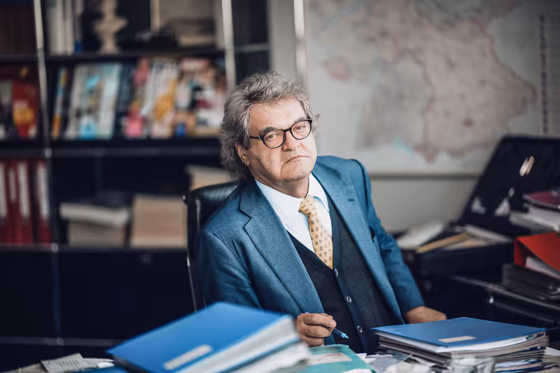Geschäftsmann mit Brille in blauem Blazer sitzt nachdenklich am Schreibtisch in modernem Büro mit Bücherregal