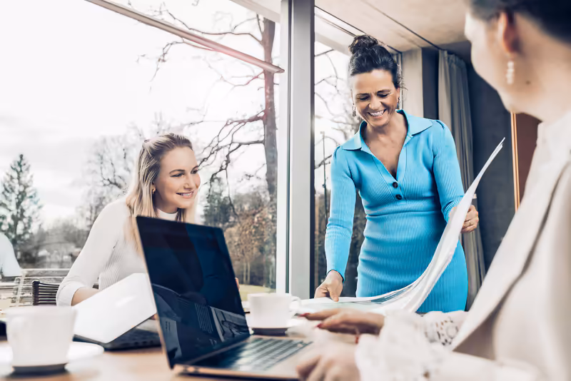 Drei Geschäftsfrauen besprechen Dokumente am Tisch in modernem Büro mit Panoramafenster und Naturblick