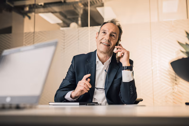 Geschäftsmann im Anzug telefoniert lächelnd am Schreibtisch im modernen Büro