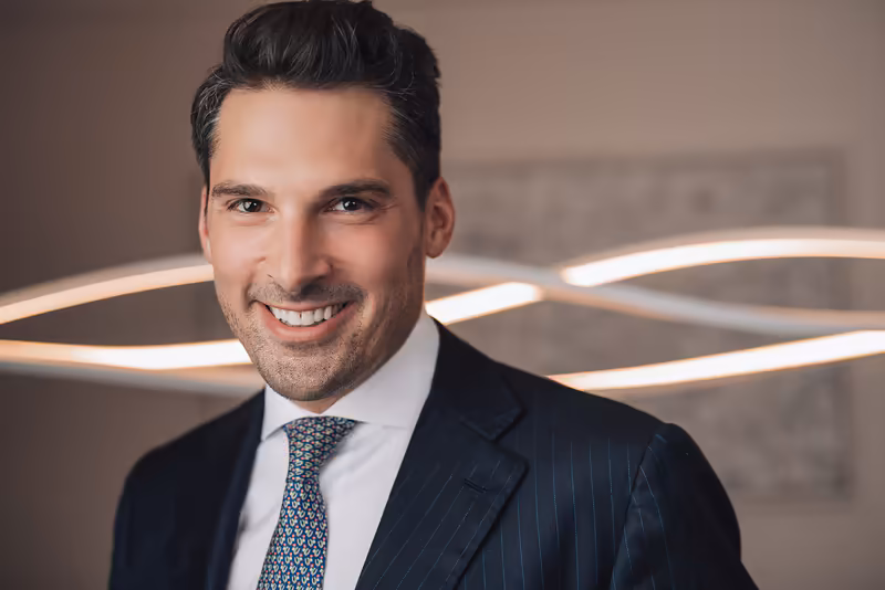 Mann im dunklen Anzug mit Krawatte lächelt vor moderner Bürokulisse mit warmen Leuchten, professionelles Business-Porträt