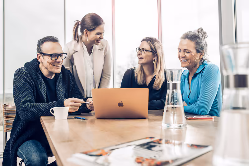 Vier Geschäftsleute im Besprechungsraum am Laptop, freundliche Stimmung, modernes Büro mit Fensterfront