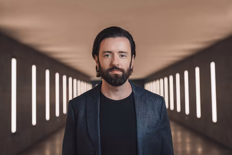 Mann mit Bart in dunklem Blazer steht in moderner Flurarchitektur mit symmetrischer LED-Beleuchtung, professionelles Portrait