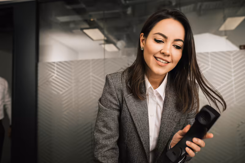 Frau in grauem Businessblazer und weißer Bluse hält Smartphone, lächelnd in modernem Büroumfeld