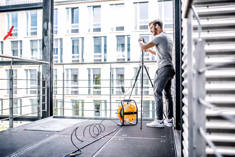 Mann mit Kamera auf urbaner Dachterrasse, gelber Rucksack, Stadtgebäude im Hintergrund