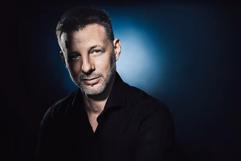 Mann in schwarzem Hemd vor blauem Hintergrund, professionelles Portrait mit direktem Blick, selbstbewusste Ausstrahlung