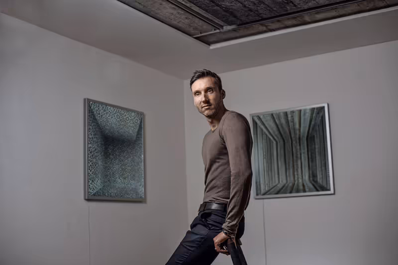 Mann in braunem Langarmshirt und dunkler Hose lehnt an Wand zwischen zwei abstrakten Kunstwerken, selbstsicherer Blick zur Kamera