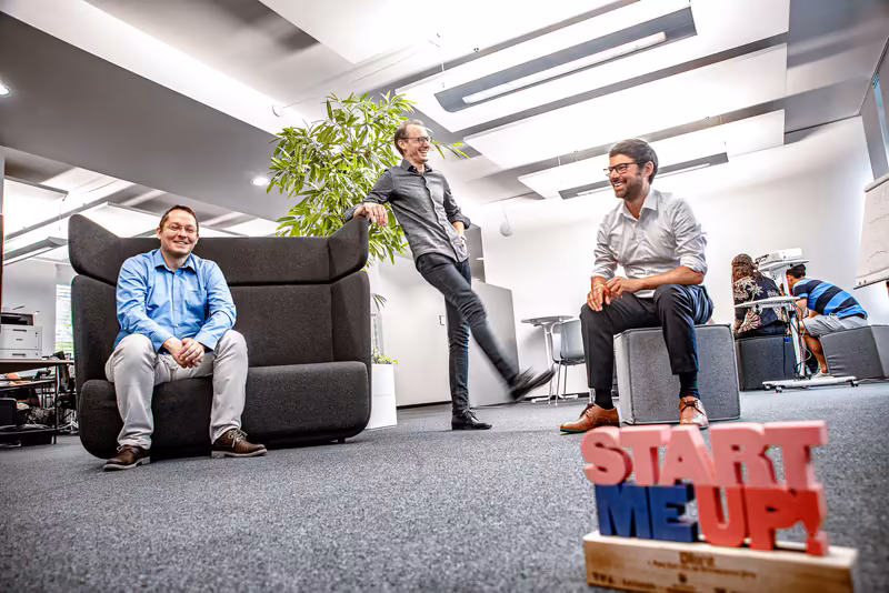 Drei Geschäftsmänner in modernem Büro mit Lounge-Bereich, entspannte Atmosphäre, Start-up-Umgebung