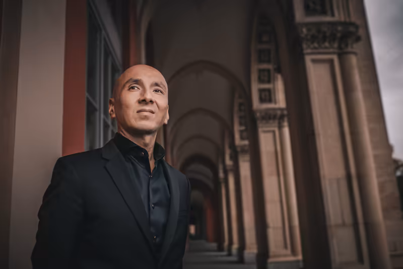 Mann in schwarzem Anzug blickt nachdenklich nach oben, klassische Säulenarchitektur im Hintergrund, professionelles Business-Portrait