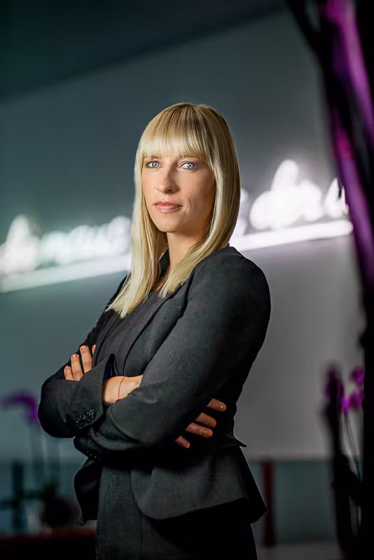 Geschäftsfrau in schwarzem Blazer, verschränkte Arme, selbstbewusste Pose vor moderner Kulisse mit Neonlicht