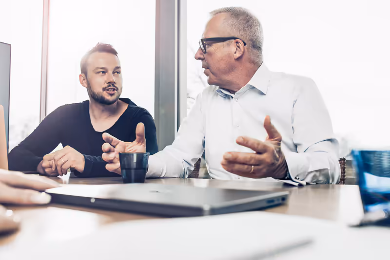 Zwei Geschäftsmänner im Gespräch am Schreibtisch mit Laptop und Dokumenten, professionelle Büroatmosphäre