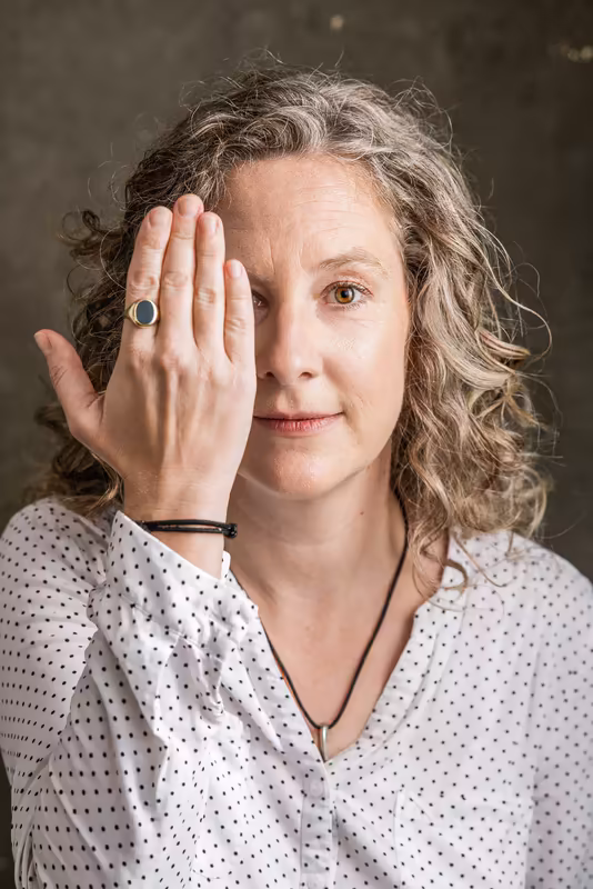 Frau mit lockigem Haar, weißem Pünktchen-Hemd und Ring hält Hand vor Gesicht, nachdenklicher Blick
