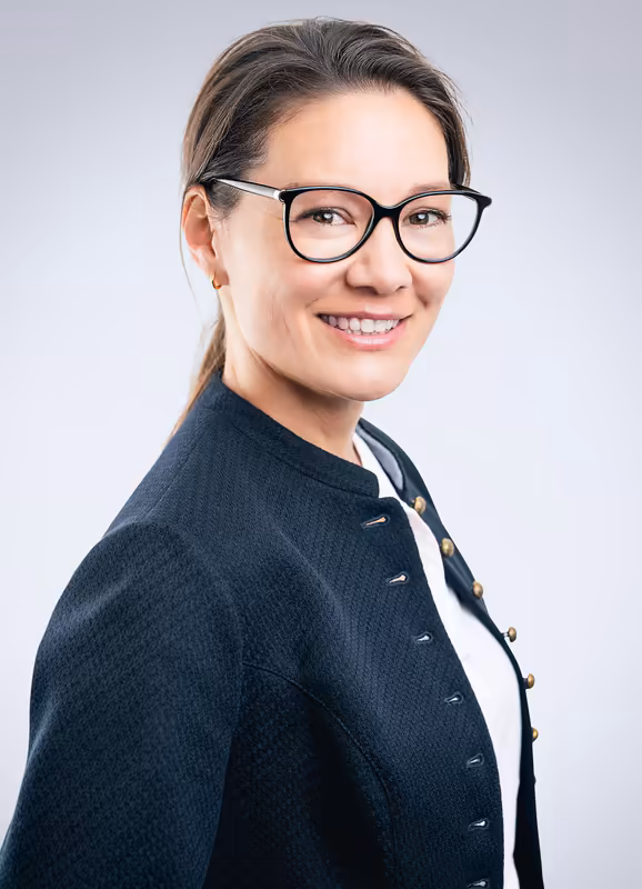 Frau mit Brille in dunklem Businessblazer lächelt zur Kamera, Porträt vor neutralem Hintergrund