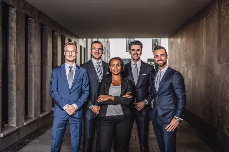 Fünf Geschäftsleute in formeller Kleidung stehen selbstbewusst in modernem Flur, Teamportrait mit professioneller Ausstrahlung