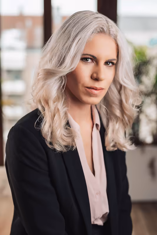 Professionelles Porträt einer Frau mit grauem Haar in schwarzem Blazer, seitlich zur Kamera, seriöser Blick, modernes Büro-Ambiente