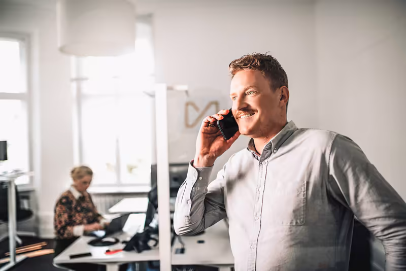 Mann im weißen Hemd telefoniert lächelnd in modernem Büro, Kollegin arbeitet am Schreibtisch im Hintergrund