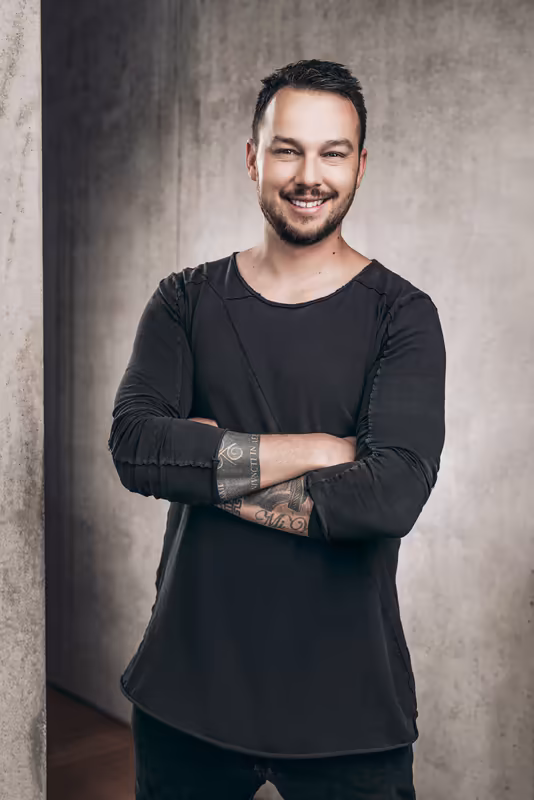 Mann mit verschränkten Armen, schwarzes Langarmshirt, lächelnd vor Betonwand, Business-Portrait