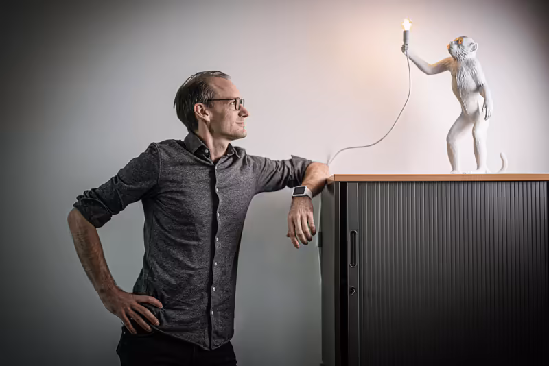 Mann in grauem Hemd lehnt an Schrank, hält Glühbirne haltender Affe an Kabel, konzeptionelle Business-Fotografie