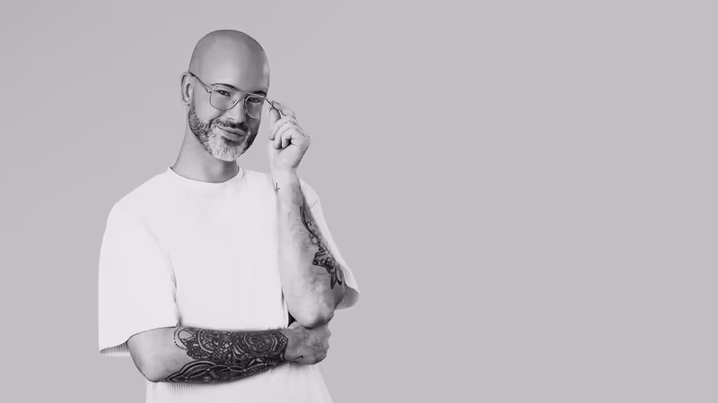 Mann mit Brille und Tätowierungen im weißen T-Shirt, selbstbewusste Pose vor grauem Hintergrund
