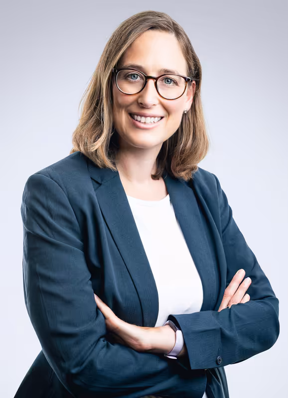 Frau mit Brille in dunkelblauem Blazer, verschränkte Arme, freundliches Lächeln, Business-Porträt vor hellgrauem Hintergrund