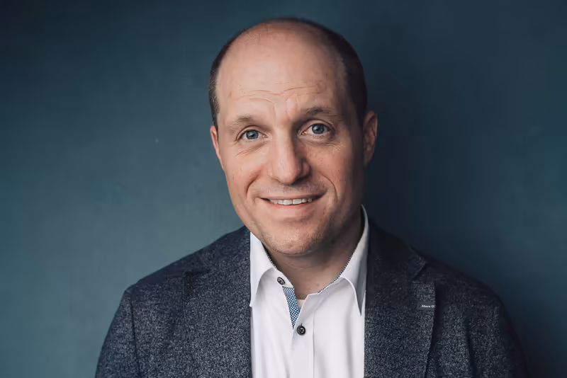 Mann mittleren Alters im Business-Portrait, lächelnd, grauer Blazer und weißes Hemd, vor graugrünem Hintergrund