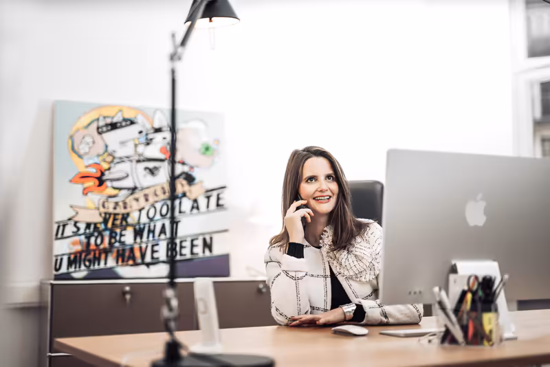 Frau am Schreibtisch mit Laptop, nachdenkliche Pose, modernes Büro mit Kunstwerk an der Wand