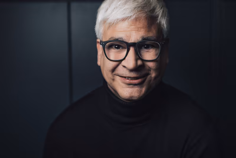 Älterer Mann mit Brille und weißem Haar, freundliches Lächeln, schwarzer Rollkragenpullover, dunkler Hintergrund, Business-Porträt