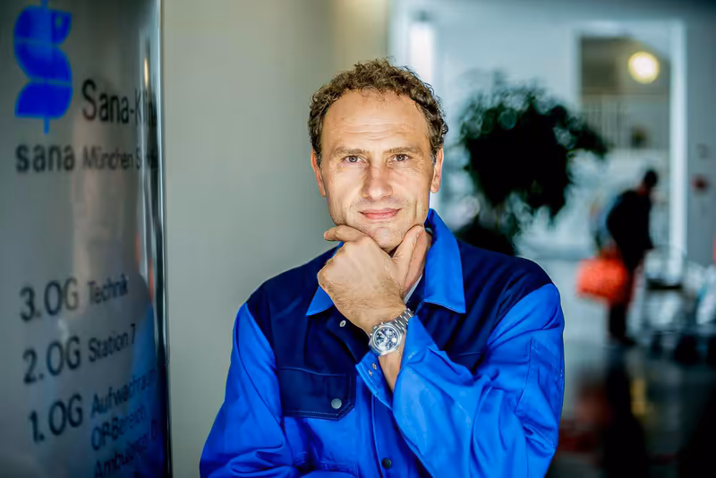 Mann in blauem Arbeitshemd, nachdenkliche Pose mit Hand am Kinn, Büroumgebung mit Schrifttafel im Hintergrund