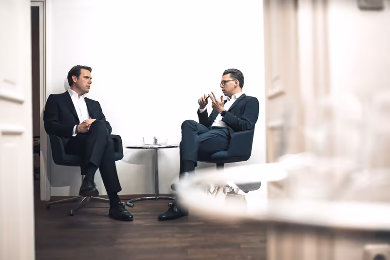 Zwei Geschäftsmänner in Anzügen führen Gespräch in modernem Büro, sitzend in Sesseln, konzentrierte Diskussion
