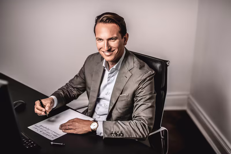 Mann im Businessanzug sitzt am Schreibtisch, schreibt auf Papier, lächelt in Kamera, professionelles Portrait