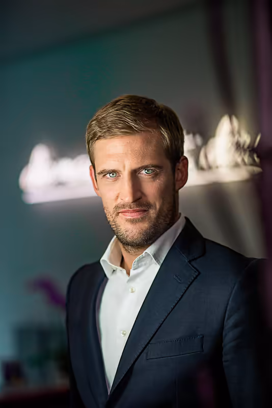 Mann im dunklen Businessblazer und weißem Hemd blickt in Kamera, professionelles Porträt vor abstraktem buntem Hintergrund