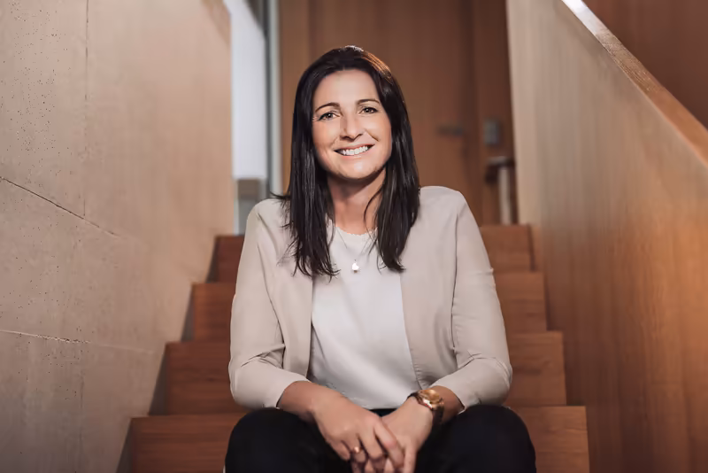 Frau sitzt lächelnd auf Holztreppe in modernem Bürogebäude, trägt helle Bluse, professionelles Portrait