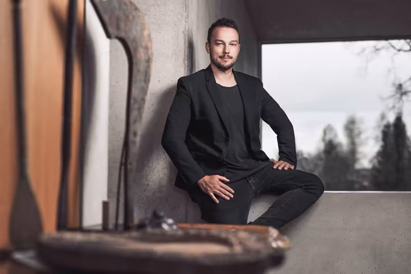 Mann in schwarzem Blazer sitzt an Betonwand, modernes Business-Portrait mit Fensterblick