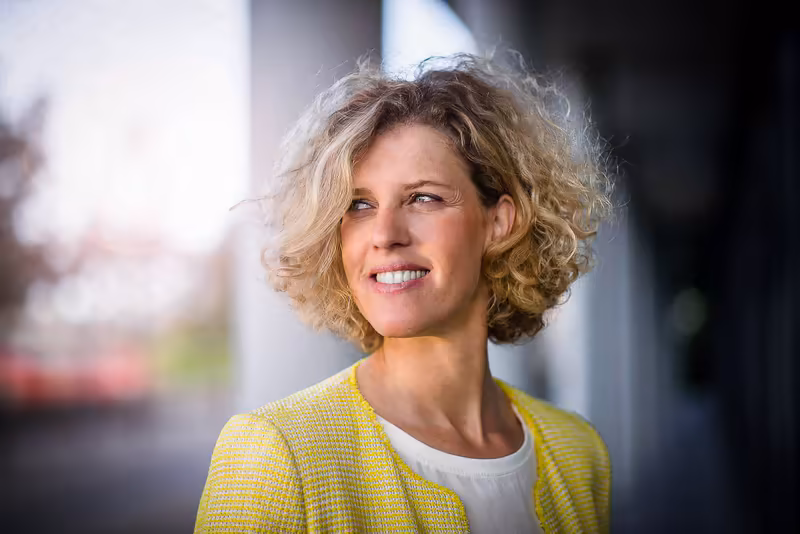 Frau mit lockigem Haar in gelbem Blazer lächelt, modernes Business-Porträt vor unscharfem Hintergrund