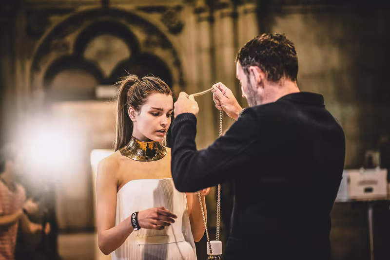 Stylist befestigt goldene Halskette an Frau in weißem Kleid in elegantem Studio mit Vintage-Architektur
