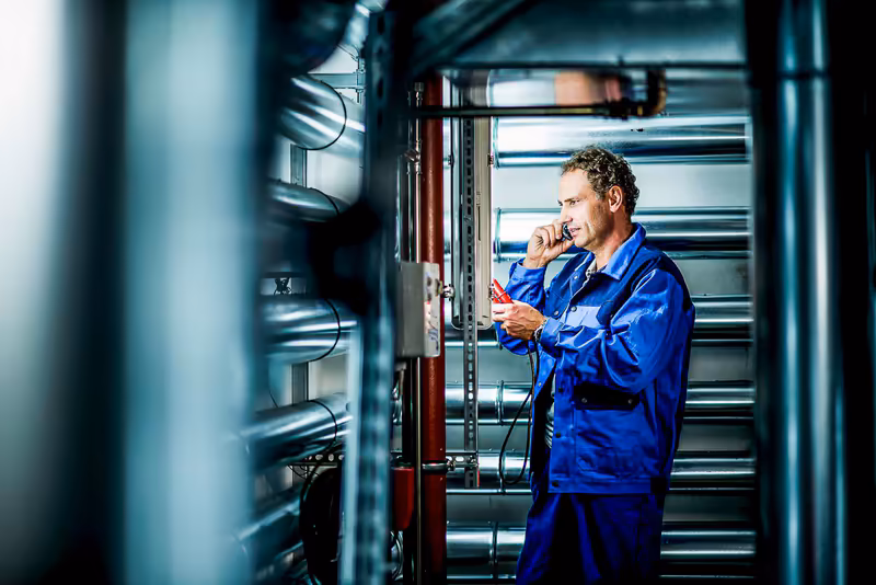 Techniker in blauem Arbeitsanzug inspiziert Industrieanlage mit Notizblock in moderner Fabrikhalle
