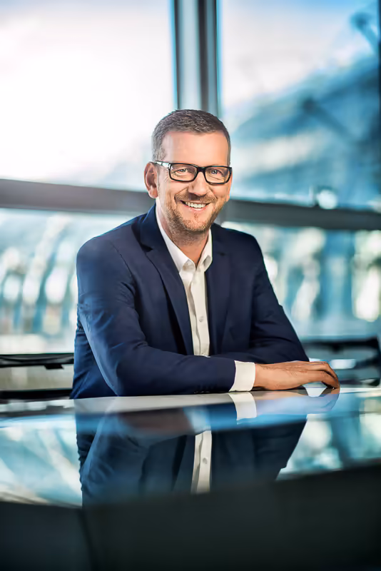 Mann in dunklem Anzug und Brille sitzt lächelnd an Glasschreibtisch vor Panoramafenster mit Stadtblick