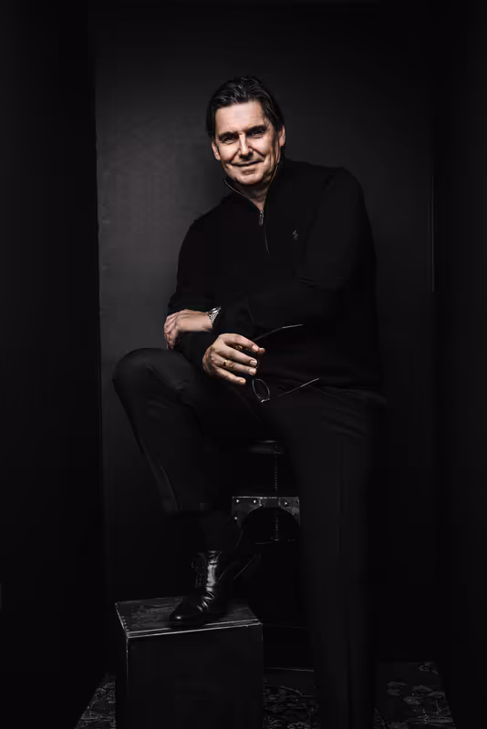 Mann in schwarzem Anzug sitzt auf Hocker, selbstbewusste Pose vor dunklem Hintergrund, Business-Portrait
