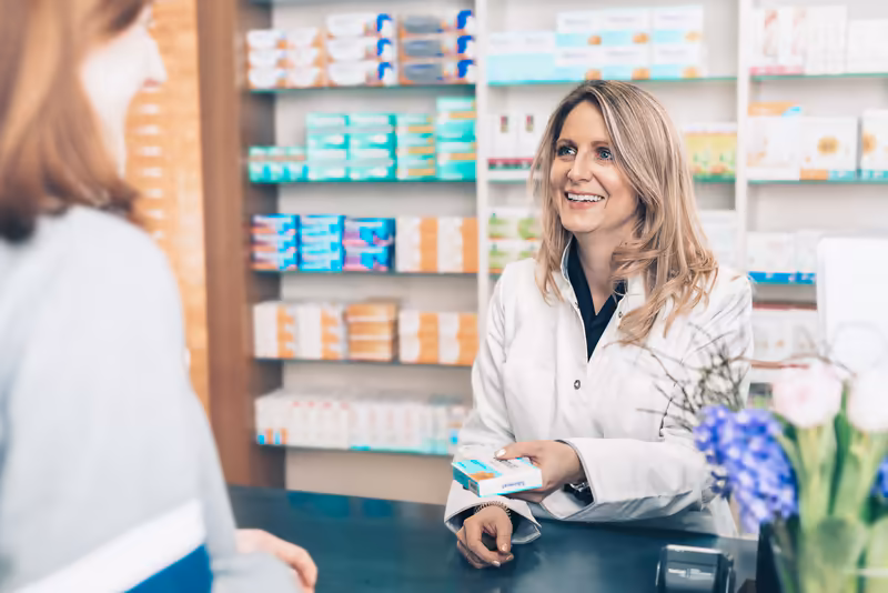 Apothekerin im weißen Kittel berät Kundin an der Apotheken-Theke, lächelnd, Medikamentenregale im Hintergrund