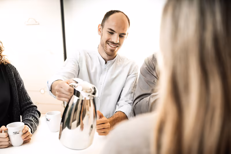 Mann in weißem Hemd schenkt Kaffee ein, lächelt, sitzt mit Kollegen am Tisch, Business-Atmosphäre