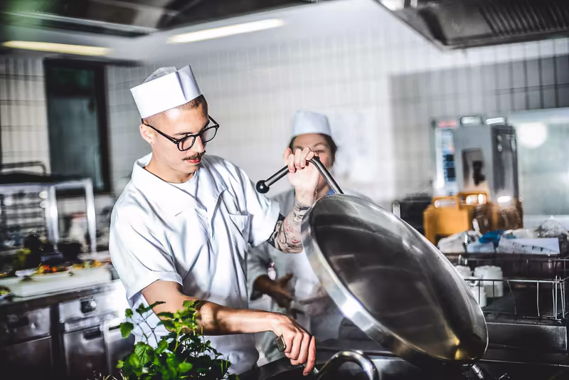 Zwei Köche in Küchenuniform arbeiten konzentriert an einer Pfanne in moderner Großküche, professionelle Kochatmosphäre