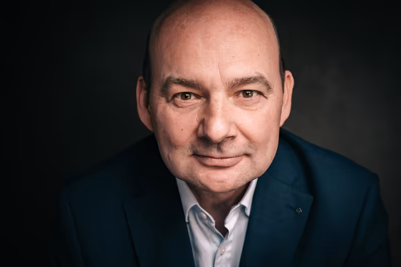Mann mittleren Alters in dunklem Businessblazer und weißem Hemd, professionelles Porträt vor dunkelgrauem Hintergrund